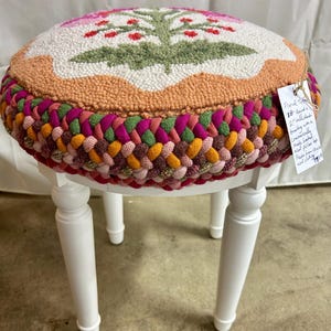 Peut inclure: Un tabouret rond avec quatre pieds blancs et une assise rembourrée présentant un motif floral crocheté en blanc, vert et rouge. Le bord de l'assise est décoré d'une épaisse bordure tressée multicolore dans des tons de rose, orange et vert. Une étiquette blanche avec du texte est attachée sur le côté.
