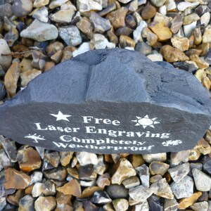 May include: A black slate rock with white text that reads "Free Laser Engraving Completely Weatherproof". The rock is surrounded by small, gray, brown, and white stones.