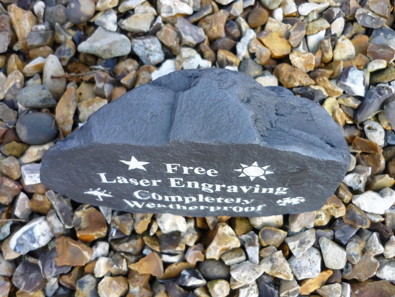 May include: A black slate rock with white text that reads "Free Laser Engraving Completely Weatherproof". The rock is surrounded by small, gray, brown, and white stones.