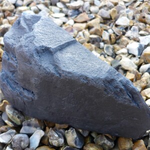 May include: A large, dark gray, irregularly shaped rock with a rough, textured surface. The rock is sitting on a bed of small, gray, brown, and white stones.