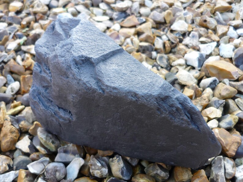 May include: A large, dark gray, irregularly shaped rock with a rough, textured surface. The rock is sitting on a bed of small, gray, brown, and white stones.