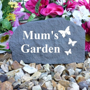 May include: A gray stone garden marker with the text "Mum's Garden" in white lettering.  The marker is surrounded by small gray and brown stones.  There are three white butterfly silhouettes on the stone.
