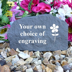 May include: A gray stone with the text "Your own choice of engraving" and a white flower design. The stone is surrounded by small gray and brown rocks.