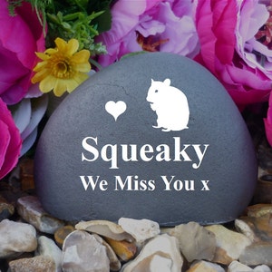 Puede incluir: Una piedra gris con un corazón blanco, una silueta blanca de un hámster y el texto "Squeaky We Miss You x". La piedra está rodeada de flores rosas.