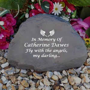 May include: A gray stone memorial with the text "In Memory of Catherine Dawes Fly with the angels, my darling..." and a pair of white angel wings.