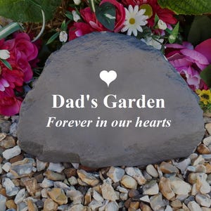 May include: A gray stone with the text "Dad's Garden Forever in our hearts" and a white heart. The stone is surrounded by pink and white flowers and small gray rocks.