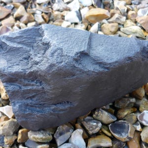 May include: A large, dark gray, irregularly shaped rock with a rough, textured surface. The rock is resting on a bed of small, light brown and gray stones.
