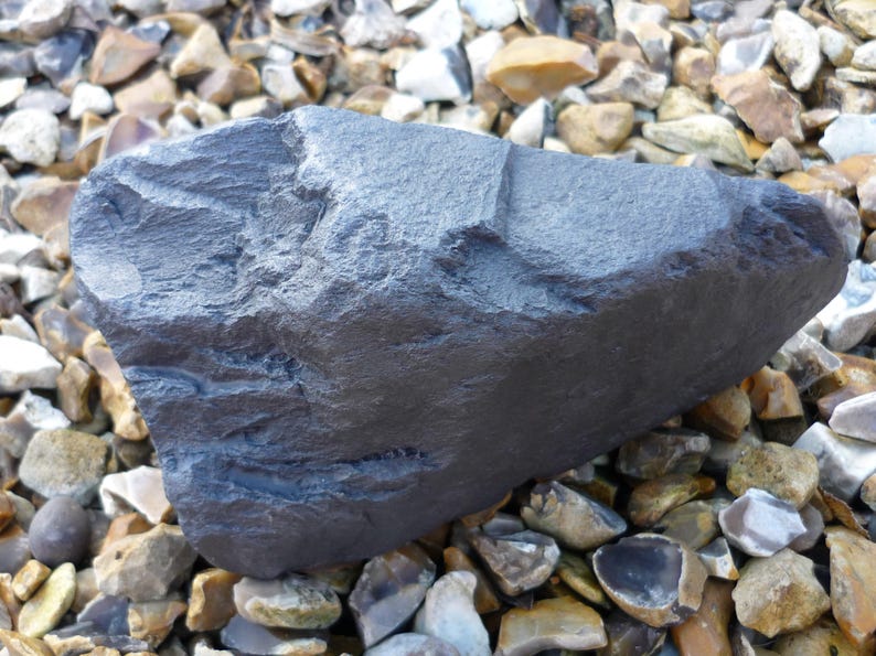 May include: A large, dark gray, irregularly shaped rock with a rough, textured surface. The rock is resting on a bed of small, light brown and gray stones.