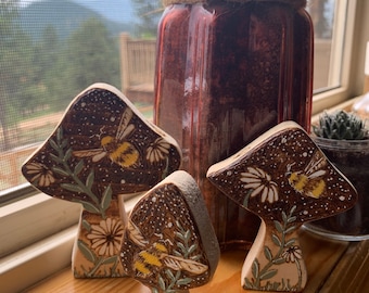May include: Three ceramic mushroom figurines with brown and white painted details. Each mushroom features a stylized bee and floral designs. The mushrooms are displayed on a wooden surface.
