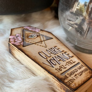 May include: A wooden coffin-shaped box with a decorative design. The box is painted with a light brown color and has a white inscription that reads "CLAIRE VOYANCE 1692-1782 SHE SHOULD HAVE SEEN IT COMING".