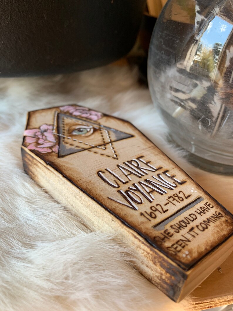May include: A wooden coffin-shaped box with a decorative design. The box is painted with a light brown color and has a white inscription that reads "CLAIRE VOYANCE 1692-1782 SHE SHOULD HAVE SEEN IT COMING".
