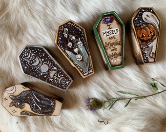 May include: Five small wooden coffins with different painted designs. One coffin has a green background with the text "I myself am strange and unusual". Another coffin has a black background with a white cat and stars. Another coffin has a black background with white moons and stars. Another coffin has a green background with white lilies. The last coffin has a black background with a white ghost holding a jack-o-lantern.
