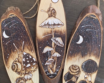 Puede incluir: Tres rodajas de madera con un fondo de madera quemada y diseños pintados en blanco y negro de setas y caracoles. Los diseños son caprichosos y presentan una luna creciente y estrellas.