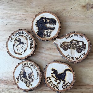 May include: Five wooden coasters with different designs and text. The coasters feature a dinosaur, a mosquito, a jeep, and the text "We spared no expense", "Life finds a way", "Hold on to your butts", and "Clever girl".