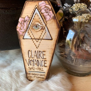 May include: A wooden coffin-shaped plaque with a silver triangle and a single eye inside. The triangle is outlined with pink flowers and a dotted line. The plaque reads "Claire Voyance 1692-1782 She should have seen it coming."