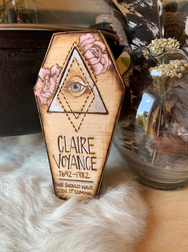 May include: A wooden coffin-shaped plaque with a silver triangle and a single eye inside. The triangle is outlined with pink flowers and a dotted line. The plaque reads "Claire Voyance 1692-1782 She should have seen it coming."