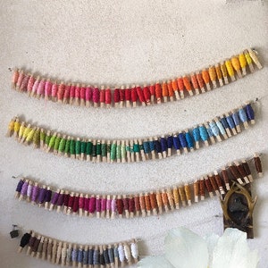 May include: A rainbow of embroidery floss spools are displayed on a white wall, each spool is held in place by a wooden clothespin.