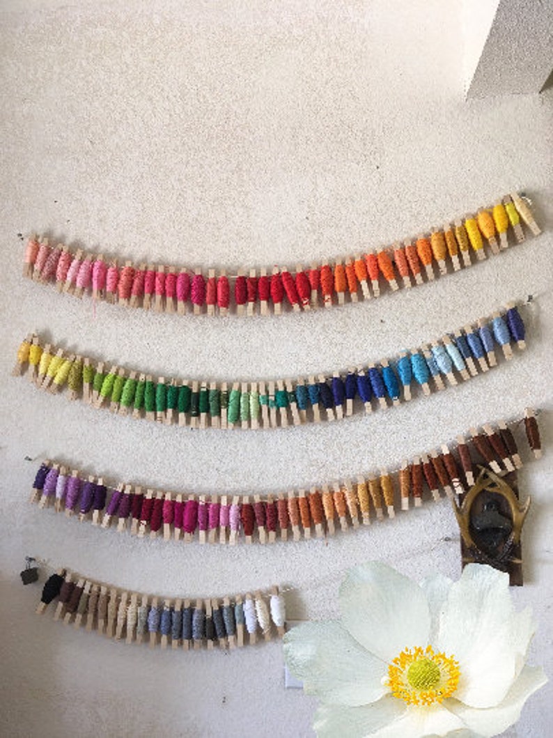 May include: A rainbow of embroidery floss spools are displayed on a white wall, each spool is held in place by a wooden clothespin.