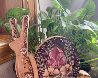 May include: A wooden snail sculpture with a pink crystal and floral design. The snail is painted with white flowers and a glittery finish. The sculpture is sitting in a brown pot with a green plant.