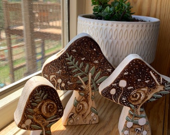 May include: Three wooden mushroom-shaped ornaments with a brown and white painted design. Each mushroom features a snail and a green leafy stem. The mushrooms are arranged in a row on a wooden surface.