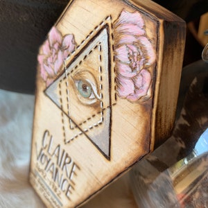 May include: A wooden plaque with a hand-painted all-seeing eye inside a triangle, surrounded by pink flowers. The text "CLAIRE VOYANCE 1912-1982" is written below the eye.