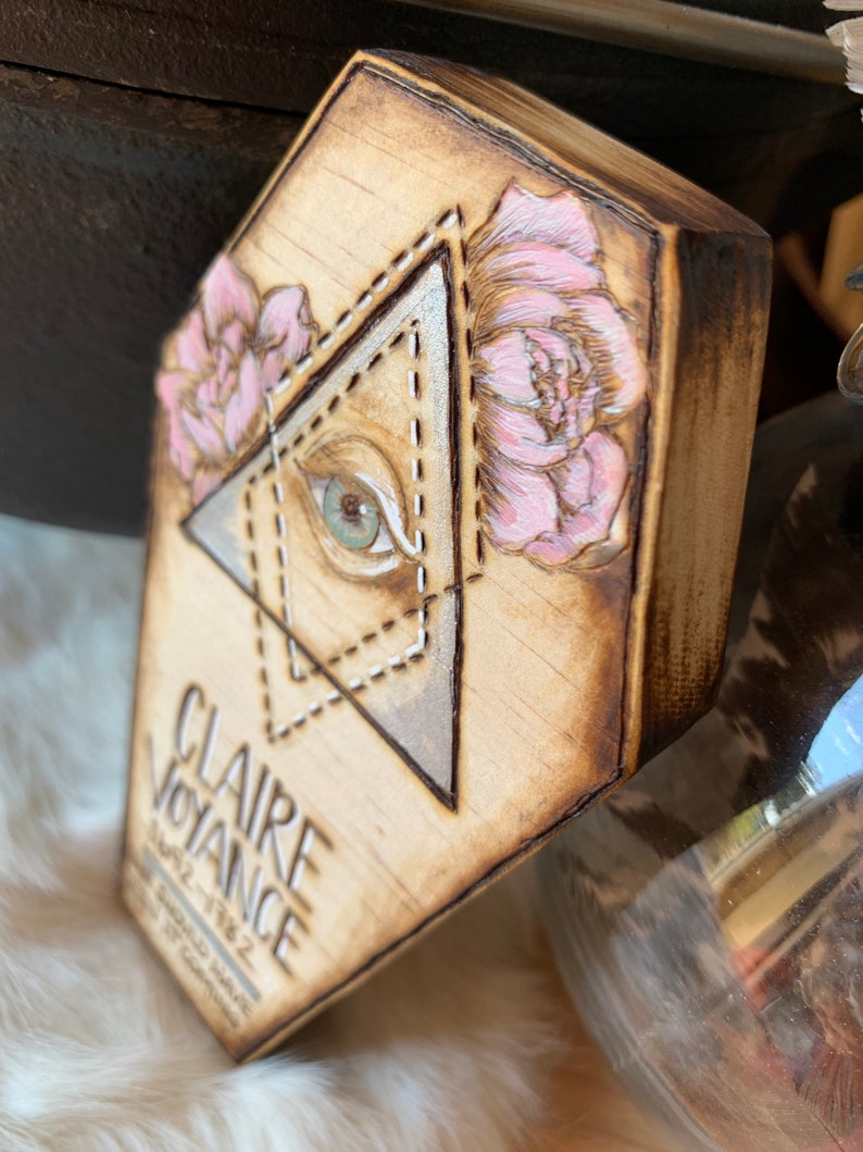 May include: A wooden plaque with a hand-painted all-seeing eye inside a triangle, surrounded by pink flowers. The text "CLAIRE VOYANCE 1912-1982" is written below the eye.