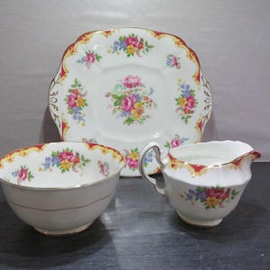 Puede incluir: Un juego de tres tazas de té vintage con estampado floral y un platillo. La taza de té y el platillo tienen un borde rojo y dorado. La taza de té tiene un estampado floral con flores rosas, rojas, amarillas y azules. El platillo tiene un estampado floral similar con un centro cuadrado.