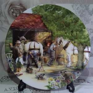 Può includere: Un piatto in ceramica bianca con una pittura colorata di un cavallo che viene ferrato in un fienile. La scena include un fabbro, un cavallo, un bambino e un gatto. Il piatto ha uno sfondo bianco e un bordo verde.