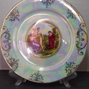 Könnte beinhalten: Ein antiker Porzellanteller mit goldfarbenem Rand und einer bemalten Szene mit drei Figuren in einem Garten. Der Teller hat eine Perlmutt-Glasur, die einen schillernden Effekt erzeugt.