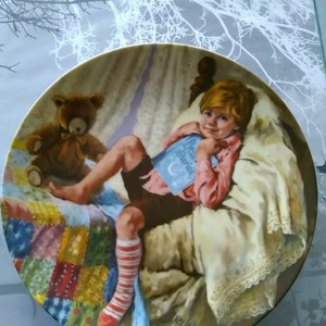 Könnte beinhalten: Ein dekorativer Keramikteller mit einem Gemälde eines jungen Jungen im Bett, der ein Buch mit dem Titel "Bedtime Stories" liest. Der Junge trägt rot-weiß gestreifte Socken und ein rotes Hemd. Ein Teddybär sitzt neben ihm im Bett. Das Bett hat eine bunte Patchwork-Decke und einen weißen Spitzenrand.