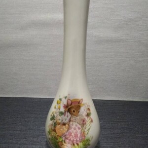 Puede incluir: Un jarrón de cerámica blanca con un borde dorado y un diseño floral con una niña con un vestido rosa y un sombrero. El jarrón mide aproximadamente 25 cm de alto.