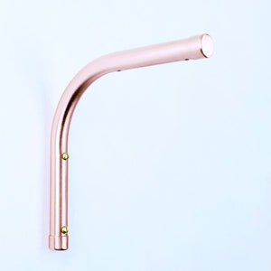 Könnte beinhalten: Eine Wandregalhalterung aus roségoldfarbenem Metall mit einem gebogenen Arm und abgerundeten Enden. Die Halterung hat zwei kleine Messing-Schrauben an der Seite.