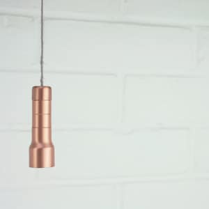 Puede incluir: Un tirador de luz de color cobre colgando de una cuerda gris. El tirador tiene un diseño cilíndrico con secciones apiladas y una base acampanada. El fondo es una pared de ladrillo blanco.
