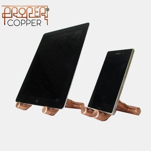 May include: Copper phone and tablet stand with a minimalist design. The stand is made of copper pipes and is designed to hold a phone or tablet at an angle for easy viewing.