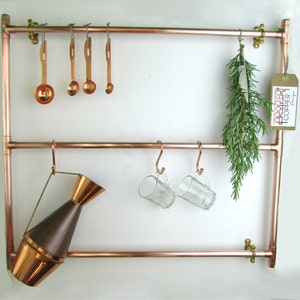 May include: Copper pipe shelf with hooks for hanging kitchen items. The shelf has two tiers and is mounted on a white wall. There are measuring spoons, mugs, a pitcher, and a bunch of rosemary hanging from the hooks.