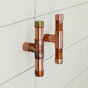 Può includere: Due ganci per asciugamani in rame con finitura rustica. I ganci hanno la forma di una T e hanno un cappuccio in rame su ciascuna estremità. I ganci sono montati su una parete piastrellata bianca.