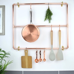 May include: Copper pipe kitchen rack with hooks for hanging pots, pans, utensils, and a cutting board. The rack is mounted on a white wall.