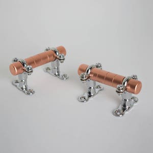 May include: Two copper drawer pulls with chrome accents. The pulls are cylindrical and have a smooth, polished finish. They are mounted on a chrome base with two screws.