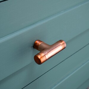 May include: A copper T-shaped drawer pull with a matte finish. The pull is mounted on a teal blue cabinet door.