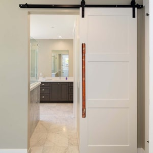 May include: A white barn door with a black sliding track and a copper door handle. The door is closed and the handle is visible.