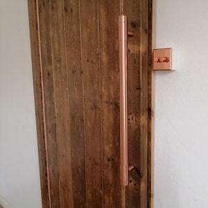 Könnte beinhalten: Ein kupferner Türgriff mit einem langen, vertikalen Design. Der Griff ist an einer Holztür mit rustikalem Finish montiert.