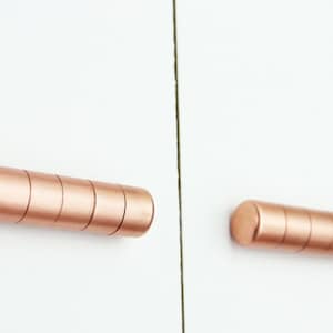 May include: Two rose gold cabinet pulls, each made of stacked round pieces of metal. The pulls are mounted on a white wall.