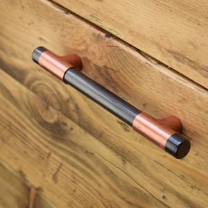 May include: Black and copper drawer pull with a rounded rectangular shape. The pull is mounted on a wooden surface.