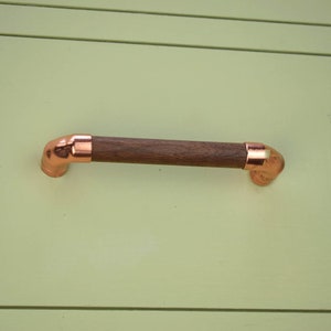 May include: A wooden drawer pull with copper end caps. The pull is a simple design with a rounded shape.