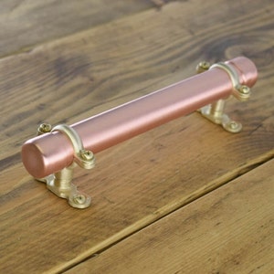 May include: A rose gold coloured metal drawer pull with gold accents. The pull is a long cylinder with rounded ends and is mounted on two gold brackets.