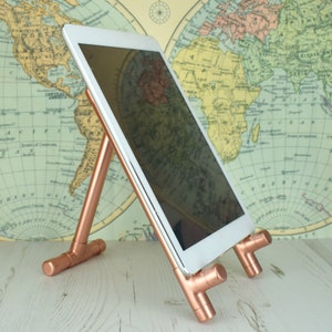 Pode incluir: Um suporte de tablet de cobre com um tablet branco apoiado nele. O suporte é feito de tubos de cobre e tem um design simples. O tablet está apoiado no suporte em ângulo, e a tela está voltada para o espectador. O suporte está sobre uma superfície de madeira branca.
