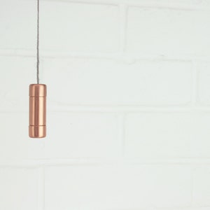 Puede incluir: Un tirador de luz cilíndrico de color cobre cuelga de un cordón gris contra una pared de ladrillos blancos. El tirador tiene un diseño simple y minimalista con un acabado metálico liso. El fondo es blanco brillante, proporcionando un fuerte contraste con el cobre.