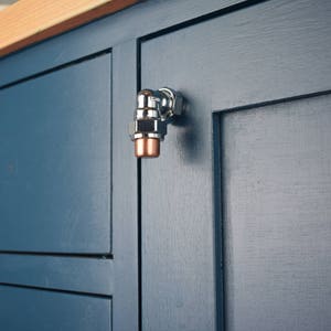 Peut inclure: Une poignée de porte de placard en chrome et cuivre en forme de raccord de tuyau. La poignée est montée sur une porte de placard bleu foncé.