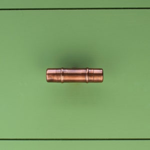 May include: A copper pipe door handle with a rounded shape. The handle is mounted on a green wooden surface.