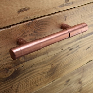 以下が含まれることがあります： 円筒形の銅色の引き出しの取っ手。取っ手は木製の表面に取り付けられています。